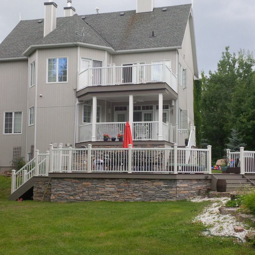 view of large grey two storey home that has a custom deck with railings and a stone wall pattern foundation